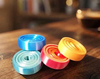 May include: Four colorful, circular objects in blue, teal, red, and yellow are arranged on a wooden surface. The objects have a layered design. Text at the bottom reads "Modèle 1 couleur à choisir dans le nuancier."