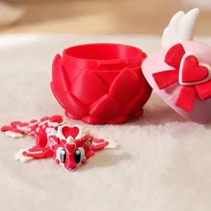 Peut inclure: Un récipient rouge en forme de cœur avec un design de lotus, un cœur rose avec des ailes et un nœud rouge, et une figurine de dragon rouge et blanc sont présentés sur une surface blanche. Le cœur a un petit cœur dessus.