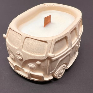 May include: A white ceramic candle shaped like a vintage Volkswagen bus. The candle has a wooden wick and is unlit.