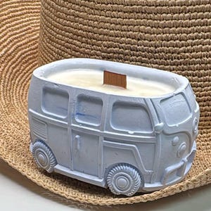 May include: A light blue candle in the shape of a vintage van. The candle has a wooden wick and sits on a woven straw hat. The van has detailed features like wheels, windows, and a front bumper. The candle is a decorative item.