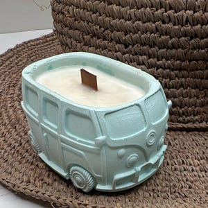 May include: A light blue candle in the shape of a vintage camper van. The candle has a wooden wick and is filled with a creamy wax. The van is detailed with wheels, windows, and a front bumper. The candle sits on a woven mat.