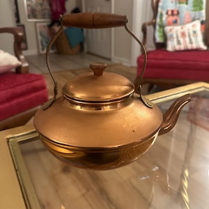 Peut inclure: Une bouilloire en cuivre avec une poign&eacute;e en bois. La bouilloire a une finition brillante et polie et un corps arrondi.