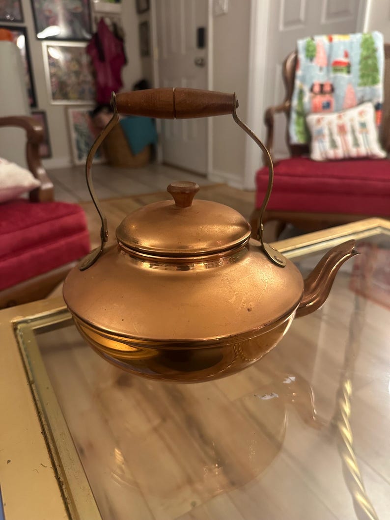 Peut inclure: Une bouilloire en cuivre avec une poign&eacute;e en bois. La bouilloire a une finition brillante et polie et un corps arrondi.