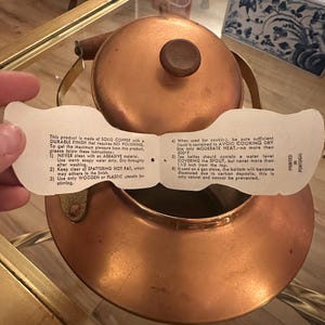 Peut inclure: Une bouilloire en cuivre avec une poign&eacute;e en bois et une &eacute;tiquette blanche avec des instructions d'utilisation. L'&eacute;tiquette indique : "Ce produit est fabriqu&eacute; en CUIVRE MASSIF avec une FINITION DURABLE qui ne n&eacute;cessite AUCUN POLISSAGE. Pour profiter au maximum de ce produit, veuillez suivre ces instructions : 1) NE JAMAIS nettoyer avec un mat&eacute;riau ABRASIF. Utilisez uniquement de l'eau savonneuse ti&egrave;de. S&eacute;chez soigneusement apr&egrave;s le lavage. 2) &Eacute;vitez les GRAISSES CHAUDES &Eacute;CLABOUSS&Eacute;ES, qui peuvent adh&eacute;rer &agrave; la finition. 3) Utilisez uniquement des ustensiles en BOIS ou en PLASTIQUE pour remuer. 4) Lorsqu'elle est utilis&eacute;e pour la cuisson, assurez-vous que suffisamment de liquide est contenu pour &Eacute;VITER DE CUISINER &Agrave; SEC. Utilisez uniquement une CHALEUR MOD&Eacute;R&Eacute;E, pas plus de 500 F. 5) Les bouilloires &agrave; th&eacute; doivent contenir un niveau d'eau COUVRANT le BEC, mais jamais plus de 1 1/2 pouce du haut. 6) Si elle est utilis&eacute;e sur une cuisini&egrave;re &agrave; gaz, le fond deviendra d&eacute;color&eacute; en raison des d&eacute;p&ocirc;ts de carbone, ce qui est tout &agrave; fait normal et ne peut &ecirc;tre &eacute;vit&eacute;."