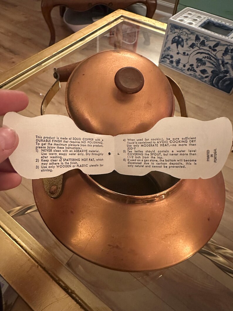 Peut inclure: Une bouilloire en cuivre avec une poign&eacute;e en bois et une &eacute;tiquette blanche avec des instructions d'utilisation. L'&eacute;tiquette indique : "Ce produit est fabriqu&eacute; en CUIVRE MASSIF avec une FINITION DURABLE qui ne n&eacute;cessite AUCUN POLISSAGE. Pour profiter au maximum de ce produit, veuillez suivre ces instructions : 1) NE JAMAIS nettoyer avec un mat&eacute;riau ABRASIF. Utilisez uniquement de l'eau savonneuse ti&egrave;de. S&eacute;chez soigneusement apr&egrave;s le lavage. 2) &Eacute;vitez les GRAISSES CHAUDES &Eacute;CLABOUSS&Eacute;ES, qui peuvent adh&eacute;rer &agrave; la finition. 3) Utilisez uniquement des ustensiles en BOIS ou en PLASTIQUE pour remuer. 4) Lorsqu'elle est utilis&eacute;e pour la cuisson, assurez-vous que suffisamment de liquide est contenu pour &Eacute;VITER DE CUISINER &Agrave; SEC. Utilisez uniquement une CHALEUR MOD&Eacute;R&Eacute;E, pas plus de 500 F. 5) Les bouilloires &agrave; th&eacute; doivent contenir un niveau d'eau COUVRANT le BEC, mais jamais plus de 1 1/2 pouce du haut. 6) Si elle est utilis&eacute;e sur une cuisini&egrave;re &agrave; gaz, le fond deviendra d&eacute;color&eacute; en raison des d&eacute;p&ocirc;ts de carbone, ce qui est tout &agrave; fait normal et ne peut &ecirc;tre &eacute;vit&eacute;."