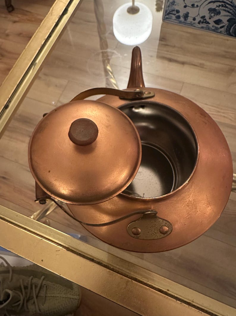 Peut inclure: Une th&eacute;i&egrave;re en cuivre avec un couvercle et une poign&eacute;e. La th&eacute;i&egrave;re est sur une table en verre avec une bordure dor&eacute;e.