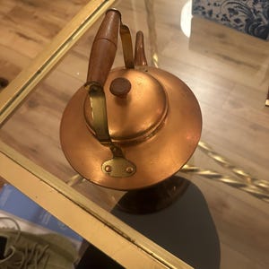 Peut inclure: Une bouilloire en cuivre avec une poign&eacute;e en bois et des accents en laiton. La bouilloire est pos&eacute;e sur une table en verre avec une bordure dor&eacute;e.