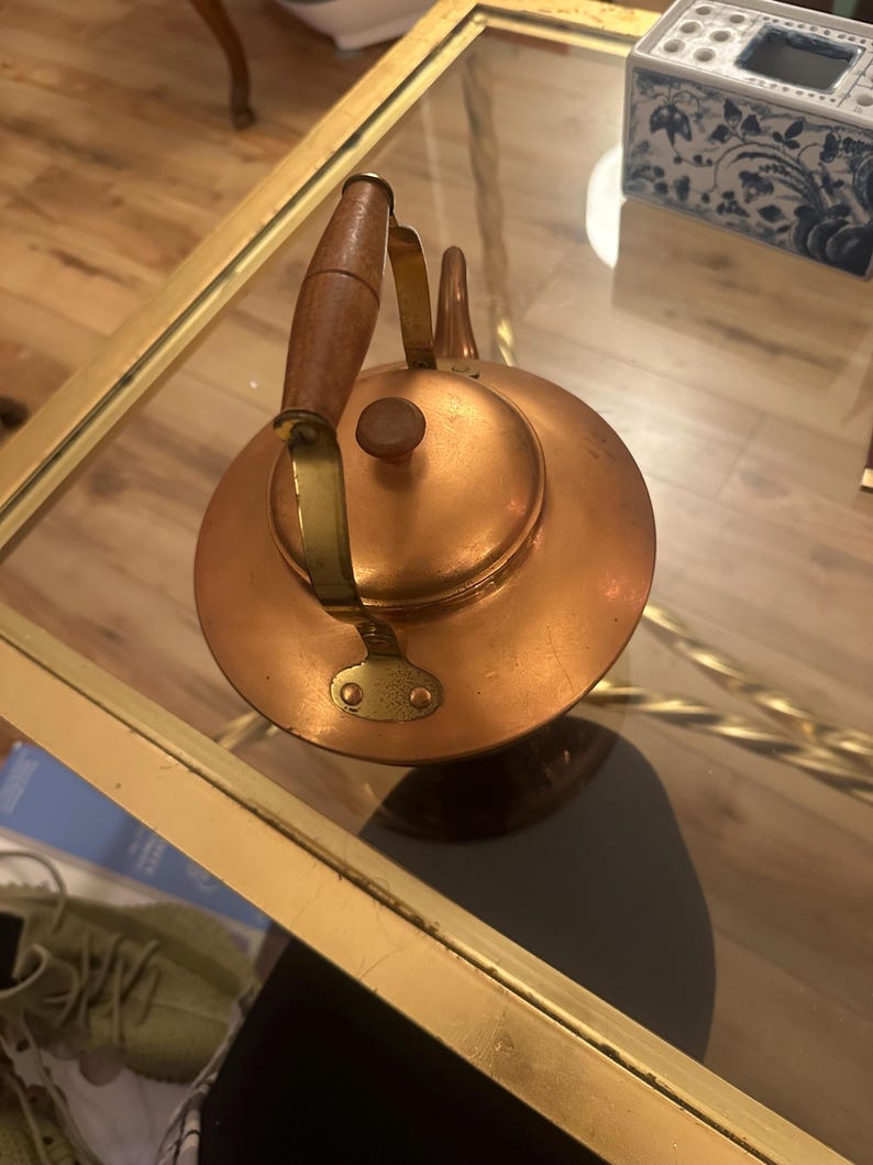 Peut inclure: Une bouilloire en cuivre avec une poign&eacute;e en bois et des accents en laiton. La bouilloire est pos&eacute;e sur une table en verre avec une bordure dor&eacute;e.