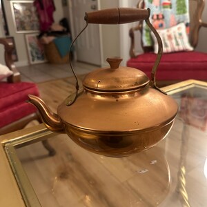 Peut inclure: Une th&eacute;i&egrave;re en cuivre avec une poign&eacute;e en bois. La th&eacute;i&egrave;re a une finition brillante et polie et est pos&eacute;e sur une table en verre.