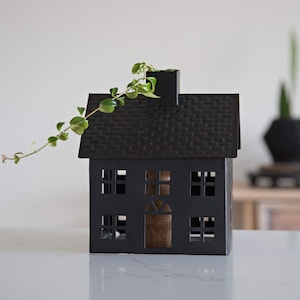 May include: Black metal house-shaped candle holder with a small plant growing out of the chimney. The house has a textured roof and cut-out windows.