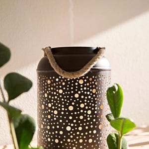 May include: A dark brown metal lantern with a rope handle. The lantern has a cylindrical shape and is covered in various sized circular cutouts. The lantern is sitting on a wooden surface with green plants in the foreground.