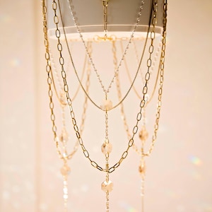 May include: An array of gold necklaces with varying chain styles and pendant designs. Some necklaces feature delicate chains with small beads, while others have larger, link-style chains. The pendants include crystal and teardrop-shaped accents.
