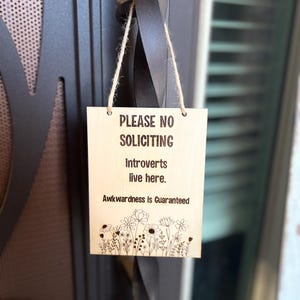 May include: A wooden sign with the text "Please No Soliciting. Introverts live here. Awkwardness is Guaranteed."  The sign is hanging on a door with a black metal handle. There are small flowers drawn at the bottom of the sign.