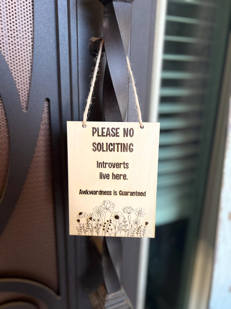 May include: A wooden sign with the text "Please No Soliciting. Introverts live here. Awkwardness is Guaranteed."  The sign is hanging on a door with a black metal handle. There are small flowers drawn at the bottom of the sign.