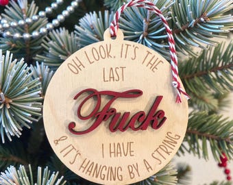 May include: A round wooden Christmas ornament with the text "Oh look, it's the last Fuck I have & it's hanging by a string." The word "Fuck" is in red script. The ornament hangs from a red and white string.