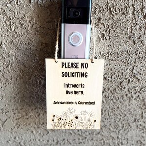 May include: A wooden sign with the text "PLEASE NO SOLICITING Introverts live here. Awkwardness is Guaranteed" and a floral design at the bottom. The sign is hanging on a wall next to a silver Ring doorbell.