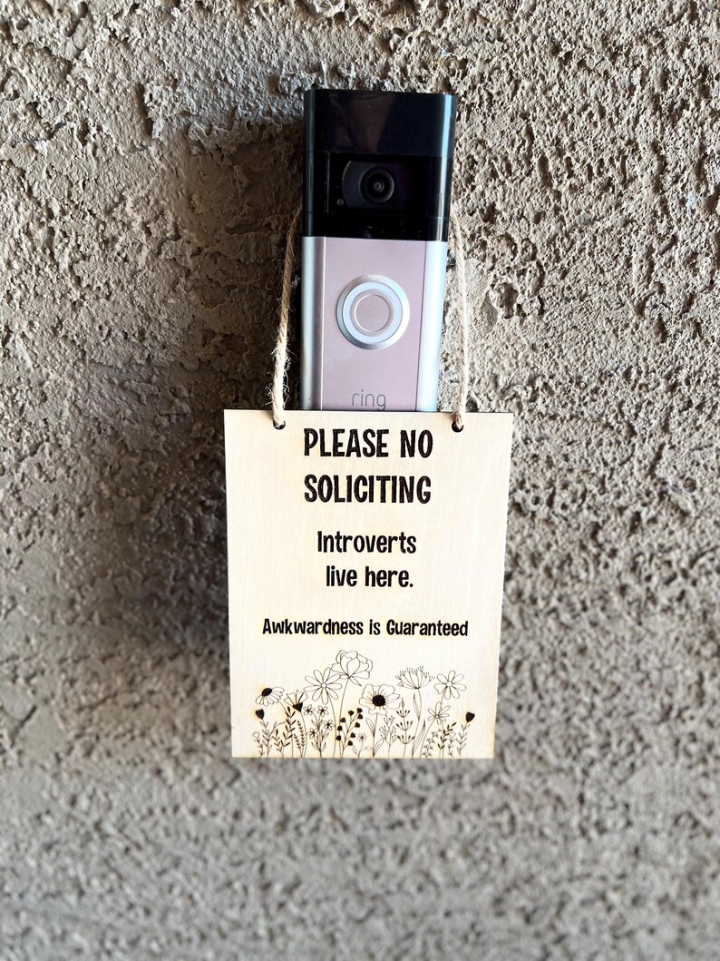 May include: A wooden sign with the text "PLEASE NO SOLICITING Introverts live here. Awkwardness is Guaranteed" and a floral design at the bottom. The sign is hanging on a wall next to a silver Ring doorbell.
