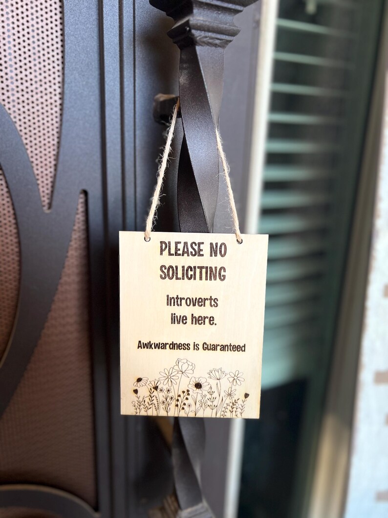 May include: A wooden sign with the text "PLEASE NO SOLICITING Introverts live here. Awkwardness is Guaranteed" and a floral design at the bottom.