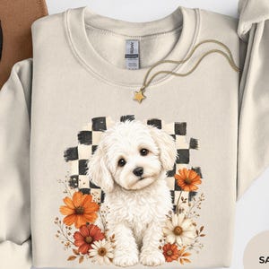 May include: A sand-coloured crewneck sweatshirt featuring a graphic of a fluffy white puppy sitting in front of a black and white checkered pattern, surrounded by orange and white flowers. A gold star pendant necklace rests on the neckline.