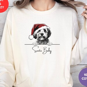May include: Ivory sweatshirt featuring a dog in a Santa hat, with the text "Santa Baby" below. The sweatshirt is labelled "Soft & Cozy" and "100% Cotton". The colour is described as "Ivory".