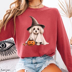 May include: A crimson long-sleeve crewneck sweatshirt featuring a graphic of a white puppy wearing a black witch hat and a ghost costume, holding a small orange jack-o'-lantern pumpkin bucket with a single piece of candy nearby.