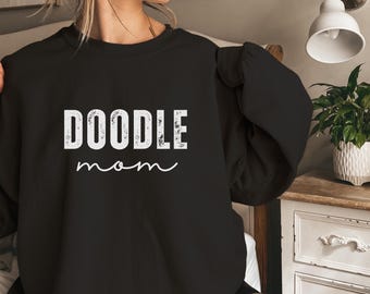 May include: Black sweatshirt featuring the text "DOODLE mom" in white. The word "DOODLE" is in a bold, distressed font, and "mom" is in a cursive script. The sweatshirt is a crew neck style.