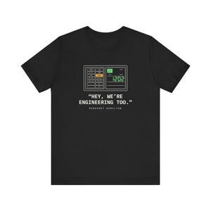 May include: A black short-sleeved t-shirt featuring a graphic of an Apollo Guidance Computer interface with the number 1202 displayed. Below the graphic is the text "HEY, WE'RE ENGINEERING TOO." and "MARGARET HAMILTON" in white.