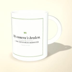 Op de afbeelding: Een witte keramische mok met een groen omrand rechthoekig kader met de tekst: My camera's broken. – THE LOCH NESS MONSTER *citation needed. De mok heeft een gebogen handvat aan de rechterkant en een glad, cilindervormig lichaam.