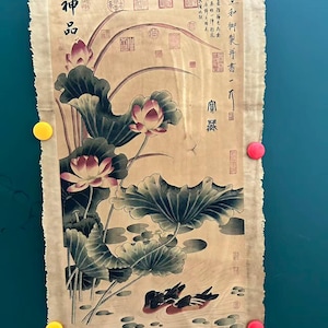 May include: An antique-style artwork featuring a lotus pond scene with ducks, lotus flowers, and large leaves. The artwork has Chinese characters and is attached to a green surface with yellow and pink magnets.