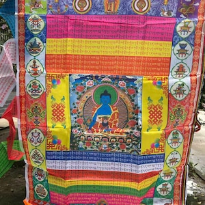 May include: A colorful prayer flag with a central image of a blue Buddha figure. The flag features various patterns, text, and symbols in red, yellow, pink, and blue. The flag is rectangular and appears to be made of fabric.