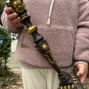Puede incluir: Objeto ritualístico detallado y ornamentado, de color bronce con detalles dorados. La parte superior presenta un rostro detallado con una corona, y la base tiene forma triangular. El objeto es sostenido por una persona con una sudadera rosa.