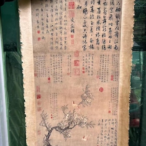 May include: An antique Chinese scroll with calligraphy and a painting of a flowering tree and birds. The artwork is on aged paper with a light brown hue, and includes red seals and intricate text. The scroll is held up by a person.