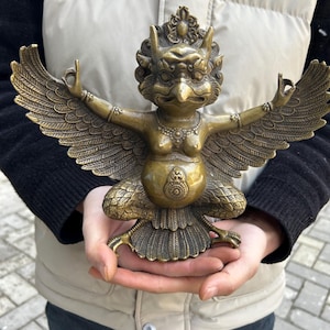 May include: A bronze-coloured statue of a mythical bird-like creature with outstretched wings and arms. The statue has intricate details, including feathers, jewellery, and a detailed head. The figure is in a seated position with its hands in a gesture.