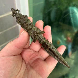 May include: Bronze-coloured, ornate dagger with a detailed handle featuring a sculpted face and intricate patterns. The blade is pointed and etched with designs. The dagger is held in a hand.