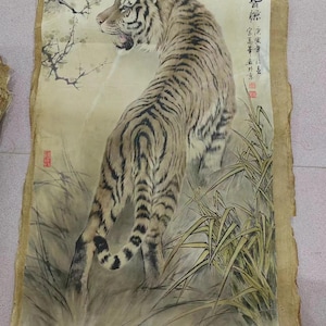 May include: An antique painting on parchment depicts a tiger with black stripes and a tan coat, standing in tall grass. The tiger is looking over its shoulder. The artwork includes Chinese calligraphy and red stamps.