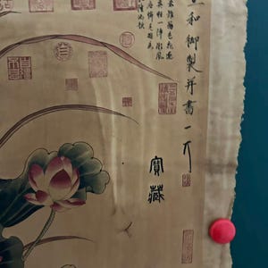 May include: A vintage scroll featuring a watercolor painting of a lotus flower with pink petals and green leaves. The artwork includes Chinese calligraphy and red seals, mounted on a beige background.