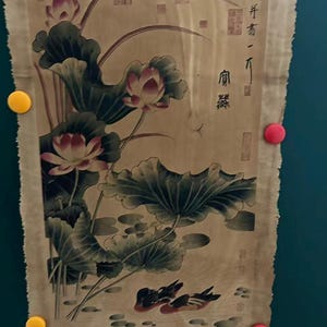 May include: An antique scroll featuring a traditional Asian painting. The artwork depicts lotus flowers with large green leaves, and two ducks swimming in a pond. The scroll has text in the upper right corner and is held in place by yellow and red magnets.