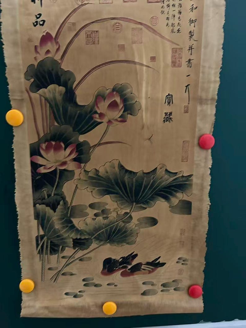 May include: An antique scroll featuring a traditional Asian painting. The artwork depicts lotus flowers with large green leaves, and two ducks swimming in a pond. The scroll has text in the upper right corner and is held in place by yellow and red magnets.