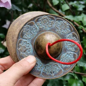 以下が含まれることがあります： アンティークブロンズ色のチベットのティンシャシンバルのペア。シンバルは、中央に真鍮色の球体があり、複雑な花柄と幾何学模様が特徴です。2つのシンバルは赤いコードでつながっています。シンバルは手で持たれています。