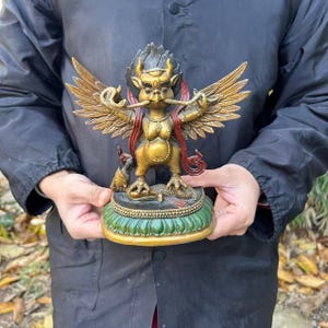 Op de afbeelding: Een bronskleurig standbeeld van een gevleugeld wezen met een vogelachtige kop, hoorns en een derde oog op het voorhoofd. Het figuur houdt een slang in de snavel, heeft rode accenten op de vleugels en staat op een groene, lotusvormige voet.