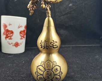 May include: A brass-colored gourd-shaped decorative item with a detailed floral design and Chinese characters. The top features a branch with leaves and a small fruit. A white ceramic cup with red illustrations is in the background.