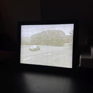 May include: A black framed picture with a grayscale image of a car driving on a winding road through a forest. The car is in the middle of the road and the trees are on either side of the road.