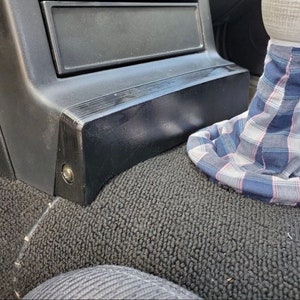 May include: A black plastic car console with a black fabric floor mat. A plaid fabric gear shift boot is visible in the foreground.