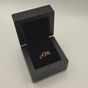 May include: A copper-coloured wire ring featuring a repeating infinity loop pattern, presented inside a square black jewellery box with a hinged lid and a black velvet interior lining that holds the ring securely in the centre of the display.