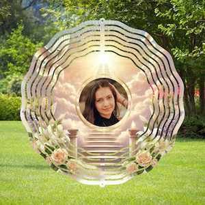 May include: A white metal wind spinner with a photo of a person in a circle in the center. The background is a cloudy sky with a stairway leading up to a bright light. There are white flowers and pink roses around the edge of the spinner.