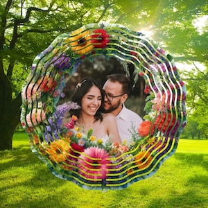 May include: A couple smiles at each other in front of a colorful floral wreath. The wreath is made of various flowers, including roses, daisies, and lilies. The couple is standing in a green field with trees in the background.