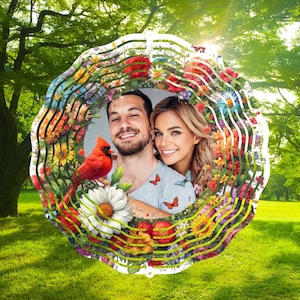 Op de afbeelding: Een witte metalen windmolen met een kleurrijk bloemenontwerp en een foto van een stel in het midden. Het ontwerp bevat een rode kardinaal, vlinders en verschillende bloemen.