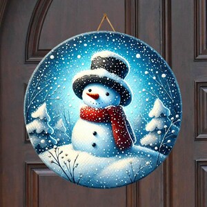 May include: A round, blue and white winter scene door hanger with a snowman wearing a black hat and red scarf. The snowman is standing in a snowy landscape with pine trees and falling snow.