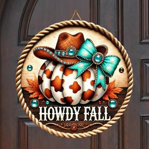 May include: A round wooden door hanger with a rope border. The centre features a brown and white cow print pumpkin with a turquoise and brown cowboy hat and bow. The text "Howdy Fall" is written in brown letters below the pumpkin.