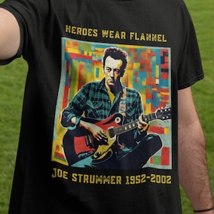 Może przedstawiać: Czarny T-shirt z kolorową grafiką przedstawiającą Joe Strummera grającego na gitarze. Grafika jest otoczona kolorowym, pikselowanym tłem. Na koszulce nadrukowany jest tekst "Heroes Wear Flannel" i "Joe Strummer 1952-2002".